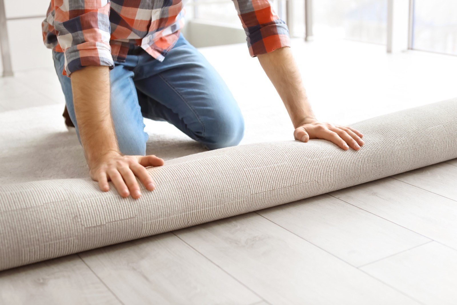 gallery-02 A man unrolls a light-colored carpet over a white wood floor, kneeling and smoothing it with his hands.